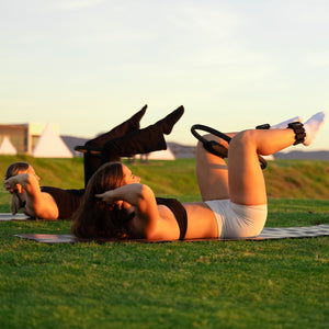 Two people exercising with yoga and pilates resistance ring on a grassy field with sunset in the background.