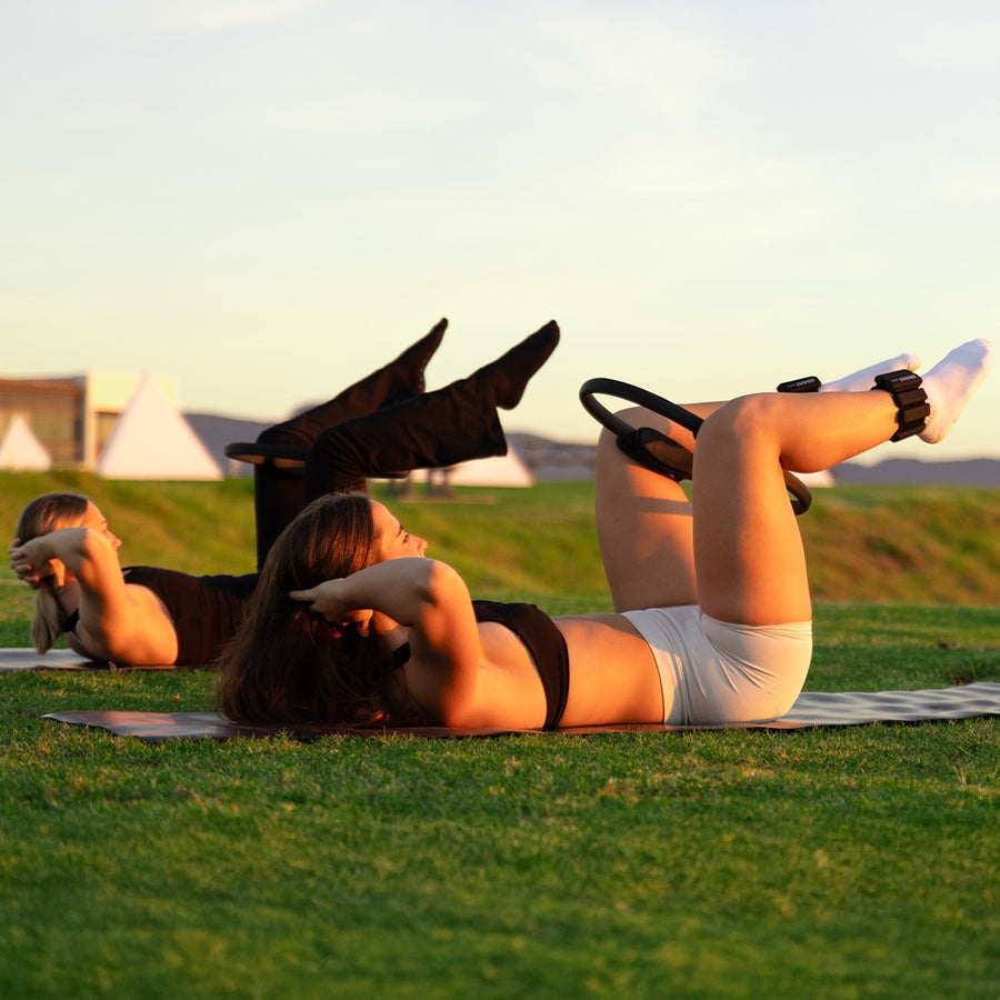 Two people exercising with yoga and pilates resistance ring on a grassy field with sunset in the background.